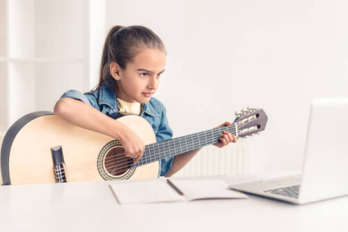Focused little kid playing acoustic guitar and watching online course on laptop while practicing at 